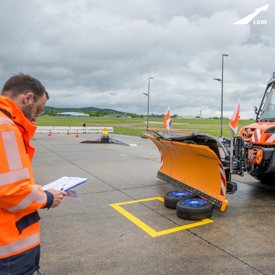 Mit einem Schneepflug werden Reifen in eine gekennzeichnete Fläche verschoben. Links daneben steht ein Mann in einer orangenen Jacke, der sich Notizen macht.