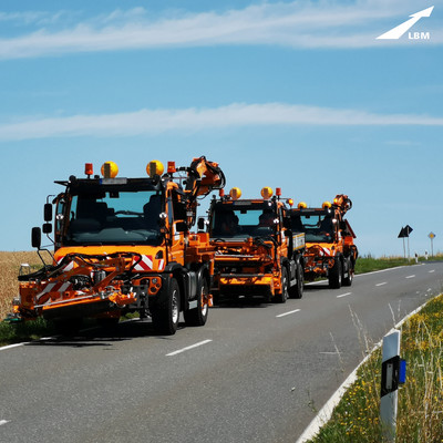 Eine Landstraße, drei orangefarbene Unimogs mit Mähaufsatz stehen hintereinander, kommen dem Betrachtenden entegegen