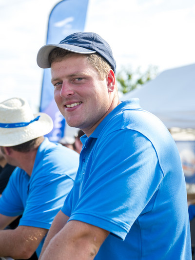 Ein Mann in blauem Polo-Shirt und mit blauer Base-Cap schaut in die Kamera und lächelt, im Hintergrund sind weitere Personen im blauen Shirt zu erahnen.