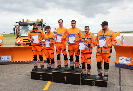 Die drei Siegerteams stehen auf einem Siegertreppchen. Im ist ein Unimog mit Schneepflug zu sehen.