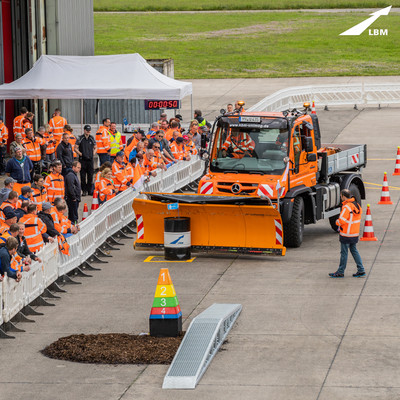 Mehrere Menschen in orangenen Jacken beobachten einen Unimog, der mit dem montierten Schneepflug ein Fass in eine gekennzeichnete Fläche verschiebt.