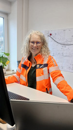 Die Ausbildungsleiterin Sonja Bernardy trägt eine orange Jacke und steht am Schreibtisch. Im Hintergrund ist ein Plan zu sehen, der an der Wand angebracht wurde. 