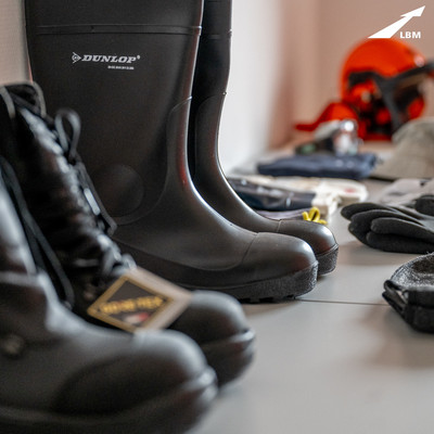 Auf einem Tisch stehen verschiedene Arbeitsschuhe, im Hintergrund sind Handschuhe und ein Helm zu erkennen