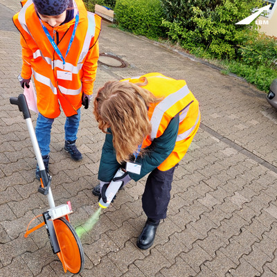 Ein Mädchen sprüht mit Farbe eine Markierung auf den gepflasterten Boden, ein Mädchen schaut dabei zu, neben ihnen ein Messrad mit dem sie vorher die Entfernung für die Markierung abgemessen haben.