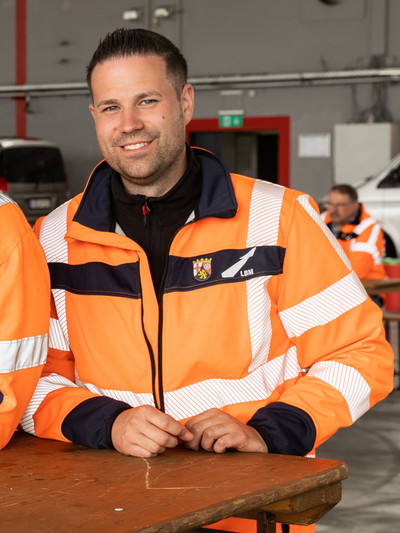 Ein Mann in orangefarbener Arbeitskleidung steht an einem Stehtisch aus Holz und schaut in die Kamera.