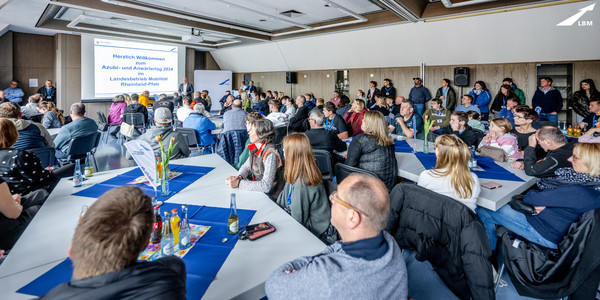 Blick in einen Raum, im Hintergrund eine Präsentationsfolie mit "Herzlich willkommen", im Raum Tische an denen viele Menschen sitzen, rechts an der Wand stehen weitere Personen