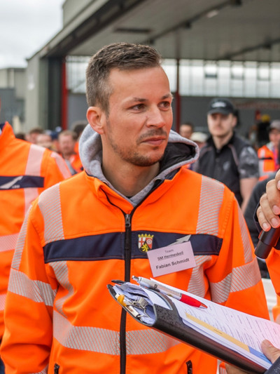 Ein Mann strahl in die Kamera. Er steht in orangener Arbeitskleidung vor einem orangenen Arbeitsfahrzeug.