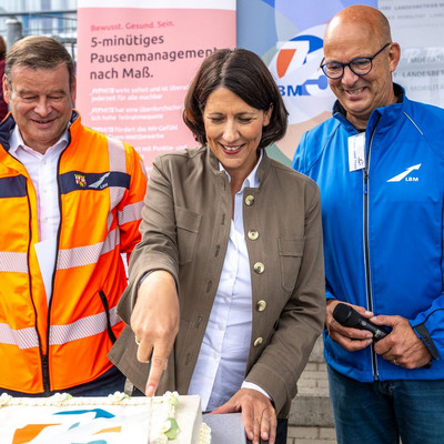 Eine Frau steht zwischen zwei Männern und schneidet einen Kuchen an.