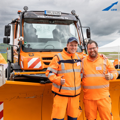 2 Männer in orangener Kleidung stehen vor einem orangenen Unimog mit montiertem Schneepflug und strahlen in die Kamera