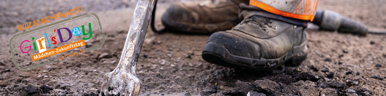 Zu sehen sind die Arbeitsschuhe eines Straßenwärters, der auf einer Straße steht. Oben links befindet sich das Logo des Girls'Day mit dem Slogan "Wir machen mit".  
