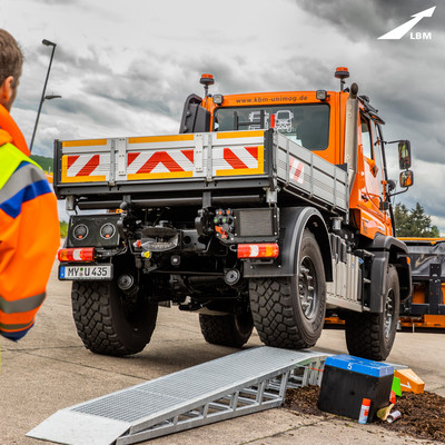 Ein Unimog mit montiertem Schneepflug fährt schräg über eine Rampe und bringt dabei eine Pyramide aus bunten Holzklötzen zu Fall.