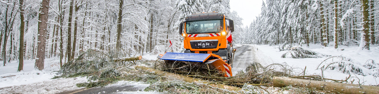 orangenes Winterdienstfahrzeug auf einer Straße im Wald, es räumt mit Schneeschild umgefallene Bäume von der Straße
