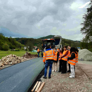 Eine Gruppe junger Mädchen steht in orangener Warnweste auf einer Baustelle, dort wird gerade eine Fahrspur asphaltiert