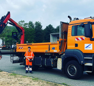Der Straßenwärter trägt Warnkleidung und steht vor einem LKW, der ebenfalls orange ist. 