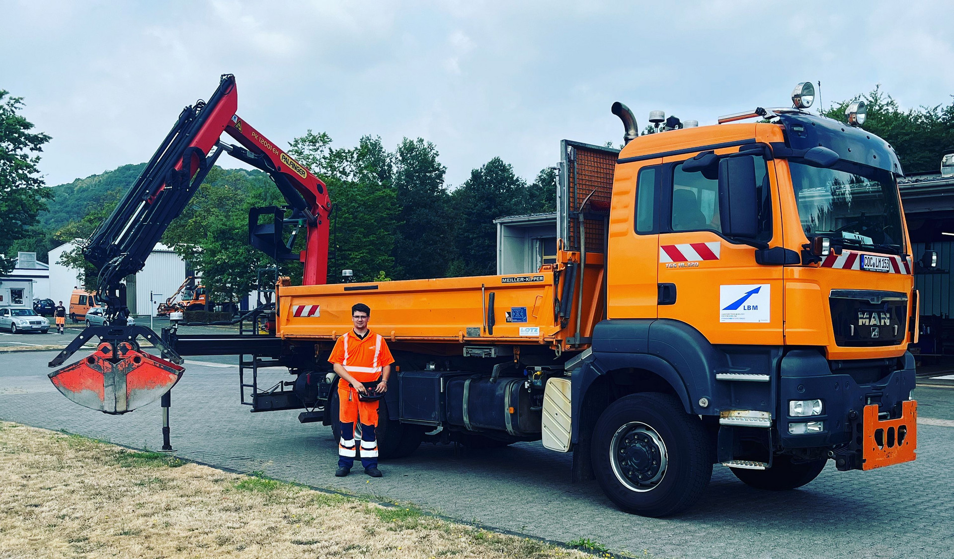 Der Straßenwärter trägt Warnkleidung und steht vor einem LKW, der ebenfalls orange ist. 