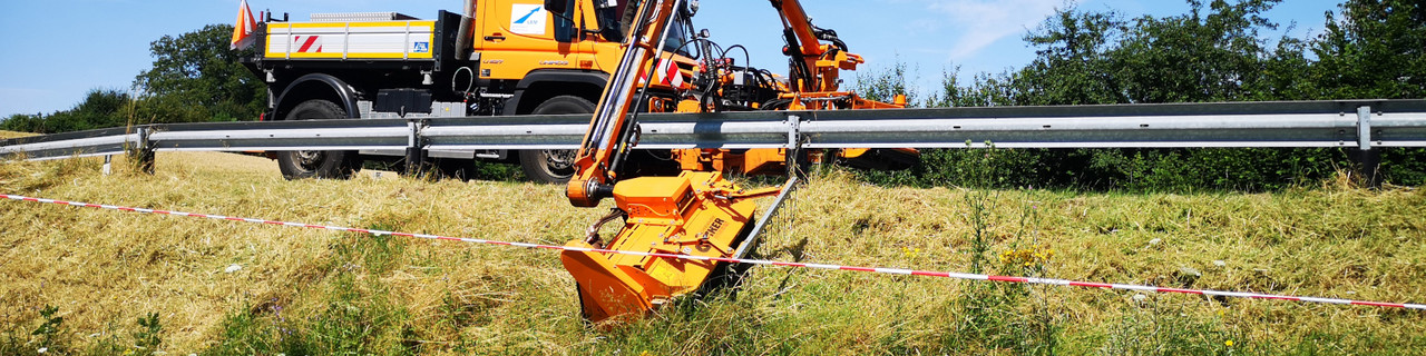 Bildausschnitt: Perspektive vom Straßenrand, Blick auf die weiter oben gelegene Straße, hinter der Leitplanke steht ein Unimog, sein Mähaufsatz ist mit einem großen Arm auf der straßenabgewandten Seite der Leitplanke und mäht die Grünfläche