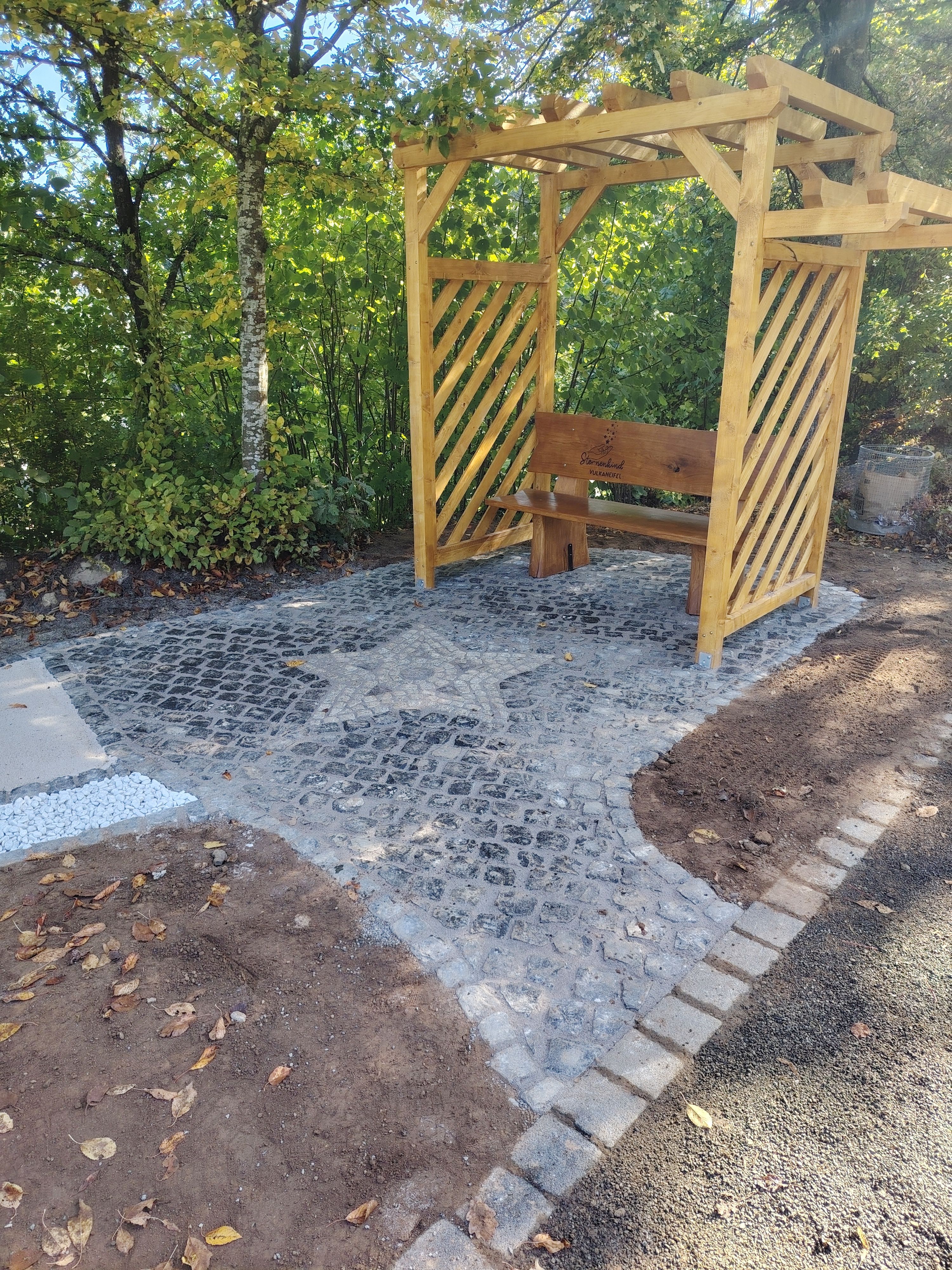 Auf einer Pflasterfläche befindet sich eine Holzbank mit der Aufschrift "Sternenkind Vulkaneifel". Die Pflastersteine wurden zudem in der Form eines Sterns angeordnet. 