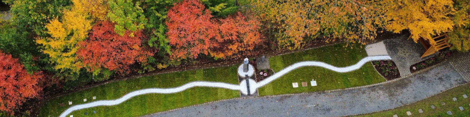 Auf dem Bild ist das fertig umgestaltete Sternenkinderfeld in Daun von oben zu sehen: eine wellenförmige Schotterfläche, eine gepflasterte Fläche in Form eines Sterns sowie Sitzmöglichkeiten und Bepflanzungen. Aufgrund der bunten Blätter der Bäume hat das Bild einen herbstlichen Charakter. 