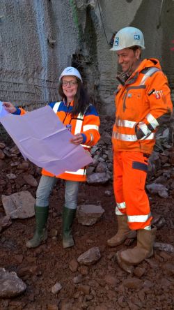 Frau Vogtmann steht mit einem Kollegen auf einer Baustelle. Sie trägt einen weißen Helm und hält einen Plan fest. Der Kollege trägt Warnkleidung.